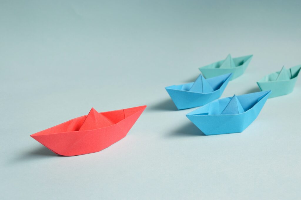 Red paper boat taking the command and leading many smaller, blue paper boats.